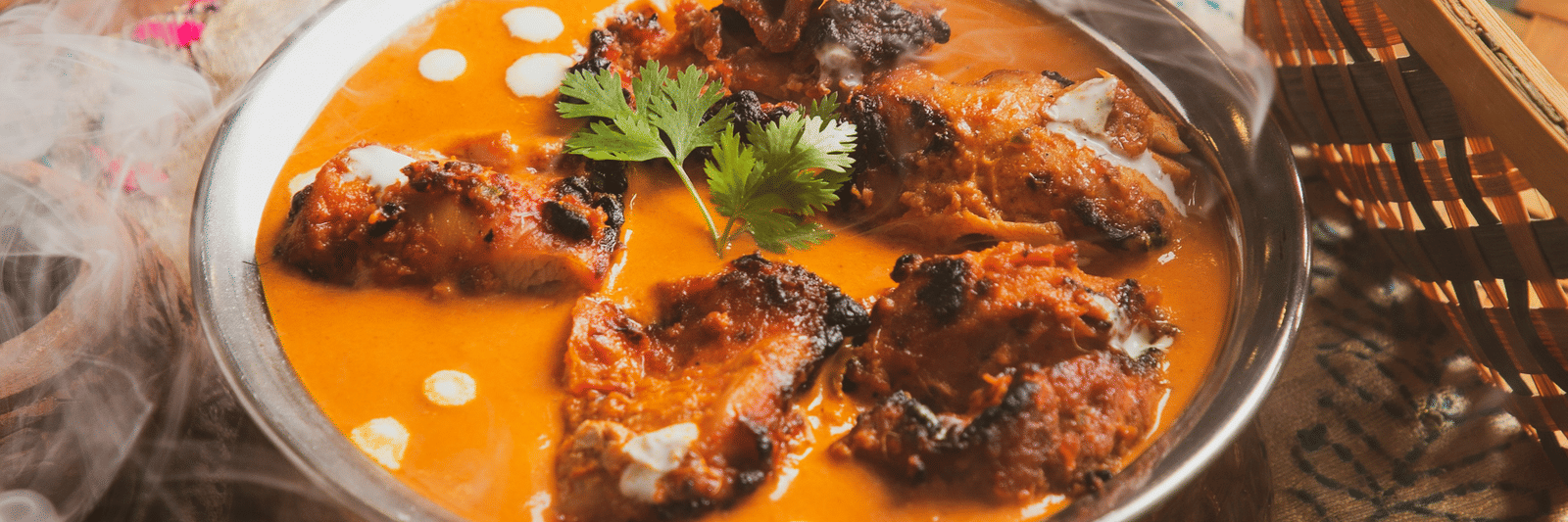 A close-up of a rich, orange-coloured curry served in a metal bowl with fresh coriander and salad.