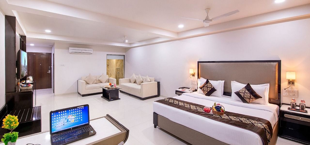 A comfortable hotel suite offers a king size bed, a separate seating area, a functional desk with a laptop, and ample natural light.