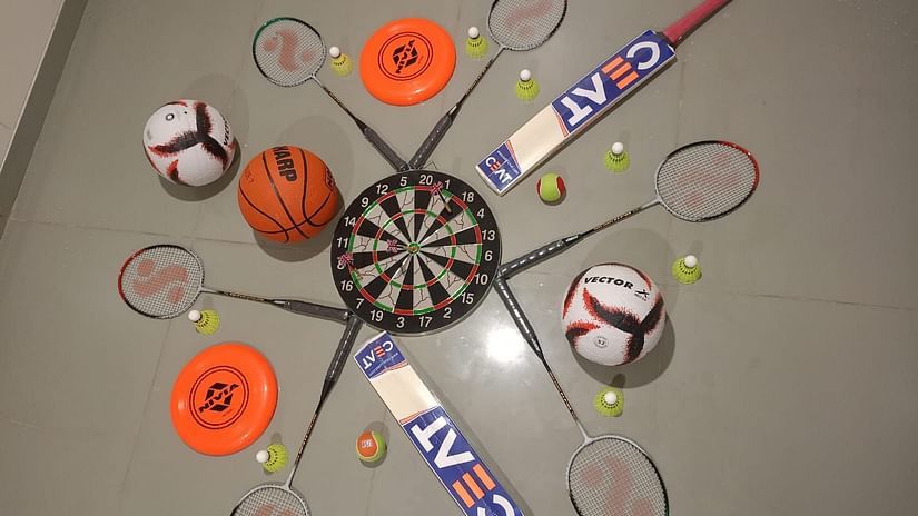 A list of sports items placed on the floor like Frisbee, cricket bat, badminton racket, basketball, football, shuttle cock, cricket ball, and a dart - Pride Resort Ambaji