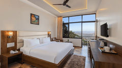 Jungle View Deluxe Room at Pride Ashiyana Resort Mussoorie featuring a plush bed facing the TV mounted on the wall and coffee maker with cups on the table below it. In the background a large window touching the floor and the roof is boasting a majestic vie
