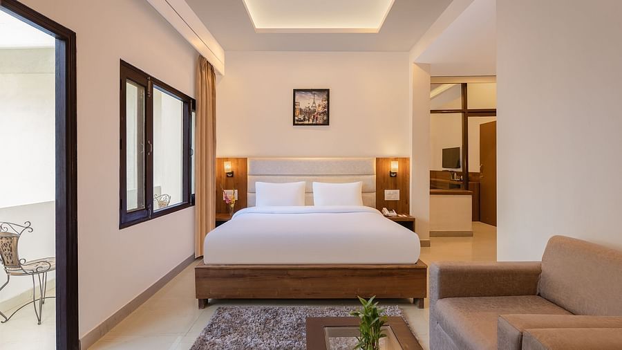 Premium Suite Room at Pride Ashiyana Resort Mussoorie featuring a plush bed placed next to the wall separating the room from balcony with a couple of lamps on both sides and a sitting space in front of it
