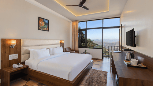 Jungle View Deluxe Room at Pride Ashiyana Resort Mussoorie featuring a plush bed facing the TV mounted on the wall and coffee maker with cups on the table below it. In the background a large window touching the floor and the roof is boasting a majestic vie