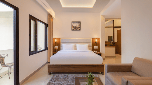 Premium Suite Room at Pride Ashiyana Resort Mussoorie featuring a plush bed placed next to the wall separating the room from balcony with a couple of lamps on both sides and a sitting space in front of it