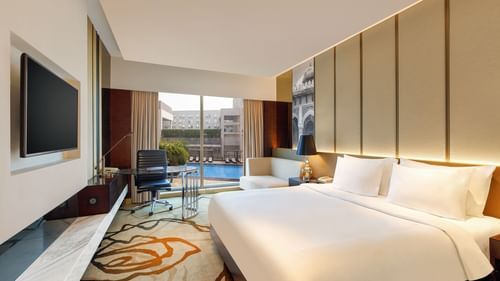 Premium King Room at Pride Plaza Hotel in New Delhi featuring a king bed in front of a TV mounted on the wall next to which a work desk is placed with a backdrop of pool view