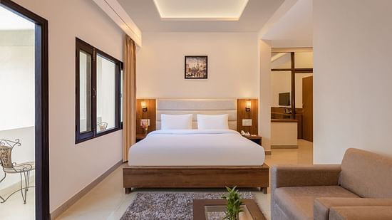 Premium Suite Room at Pride Ashiyana Resort Mussoorie featuring a plush bed placed next to the wall separating the room from balcony with a couple of lamps on both sides and a sitting space in front of it