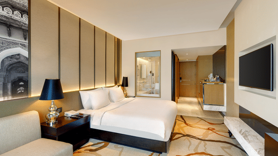 A bedroom at Pride Plaza Hotel, New Delhi featuring a bed, a sofa, bedside table with lamp on either side, and a flat TV screen.