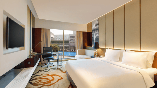 Premium King Room at Pride Plaza Hotel in New Delhi featuring a king bed in front of a TV mounted on the wall next to which a work desk is placed with a backdrop of pool view