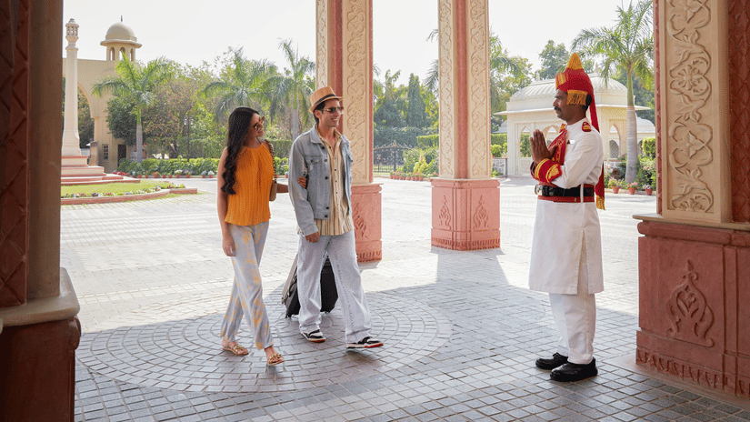 A hotel entrance at Heritage Village Resorts & Spa, Manesar, with a uniformed doorman standing near two guests walking through a covered porch.