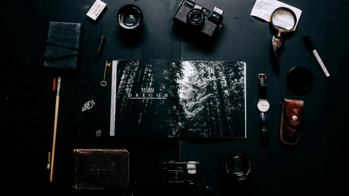 Flat lay of photography and travel essentials, including a vintage camera, watch, magnifying glass, notebook, key, and an open magazine featuring forest imagery, all arranged on a dark surface.