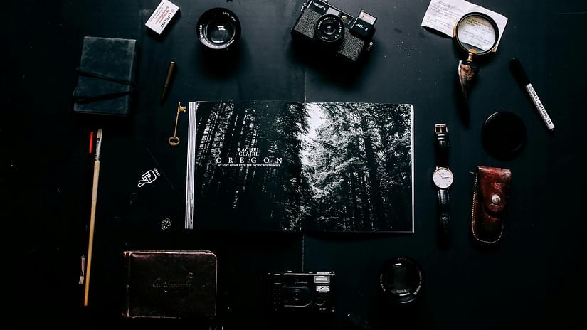 Flat lay of photography and travel essentials, including a vintage camera, watch, magnifying glass, notebook, key, and an open magazine featuring forest imagery, all arranged on a dark surface.