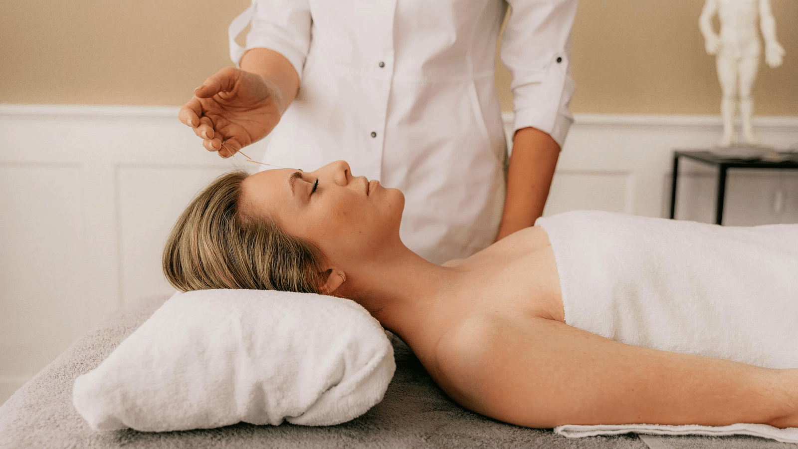 A woman lying on her back in a white towel getting acupuncture treatment in a spa.