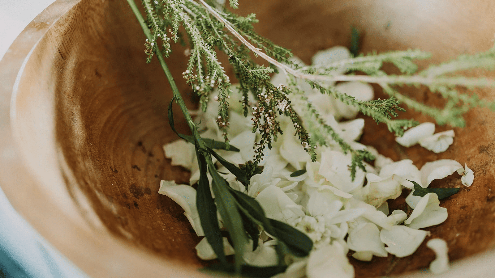 White flower petals and some green herbs placed inside a wooden bowl.