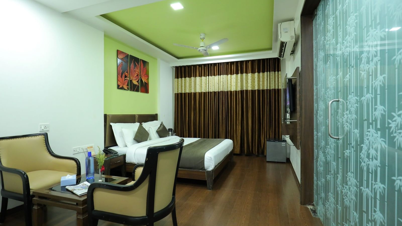 A bedroom at Krushnai Resort with a bed, 2 chairs, a glass parting on one side, and a vibrant green ceiling.