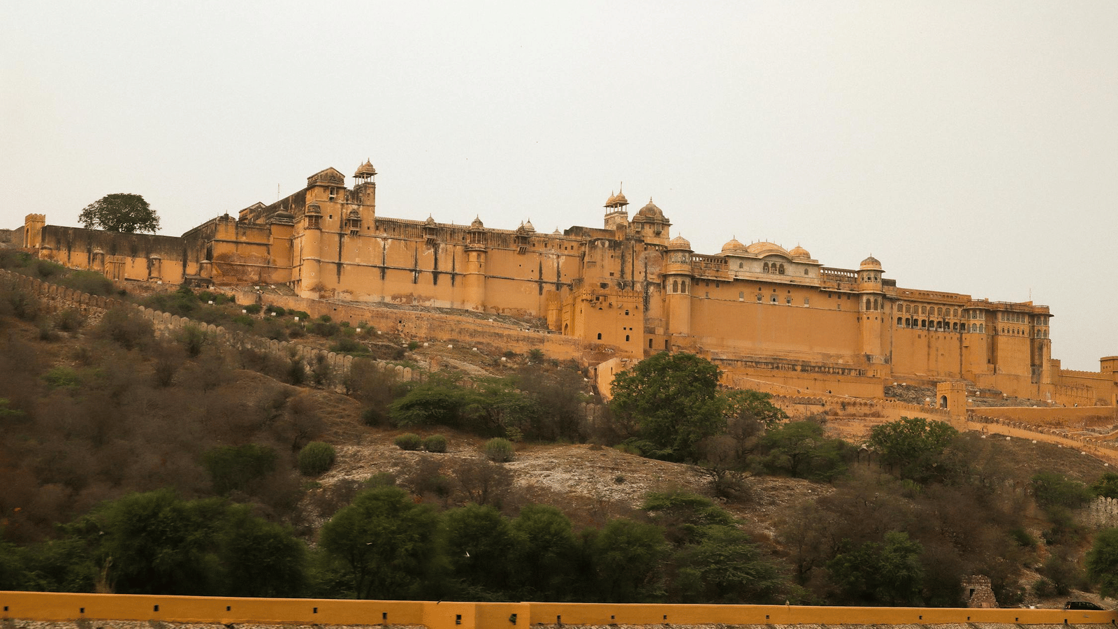 A sprawling fort complex with towers and walls extends across a hilly landscape under a hazy sky.