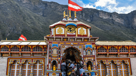 A full frontal view of the Badrinath Temple featuring its iconic multi-coloured architecture and golden spires, set against a backdrop of steep, misty Himalayan mountains.