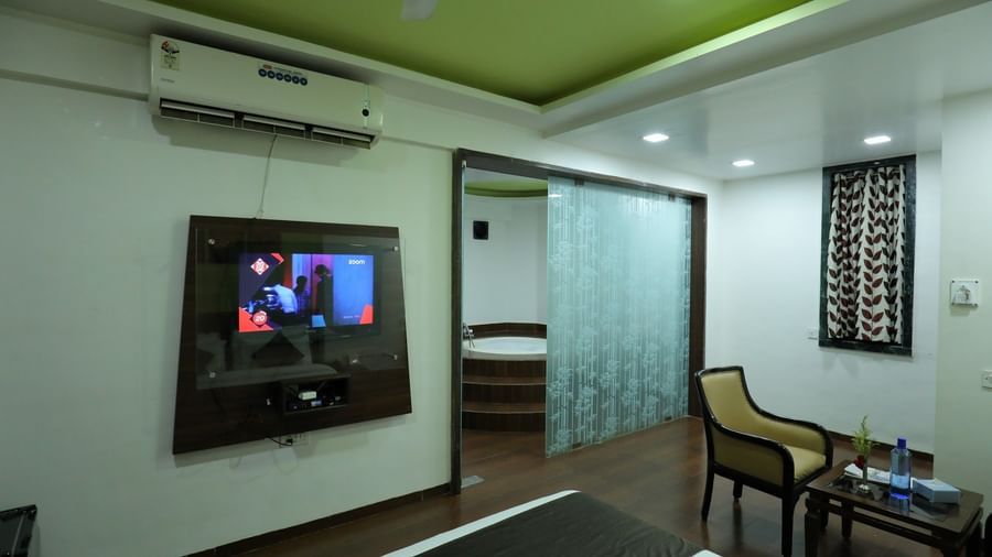 A wide view of a Krushnai Resort bedroom with a wall-mounted TV, seating area, and a glimpse of the washroom.