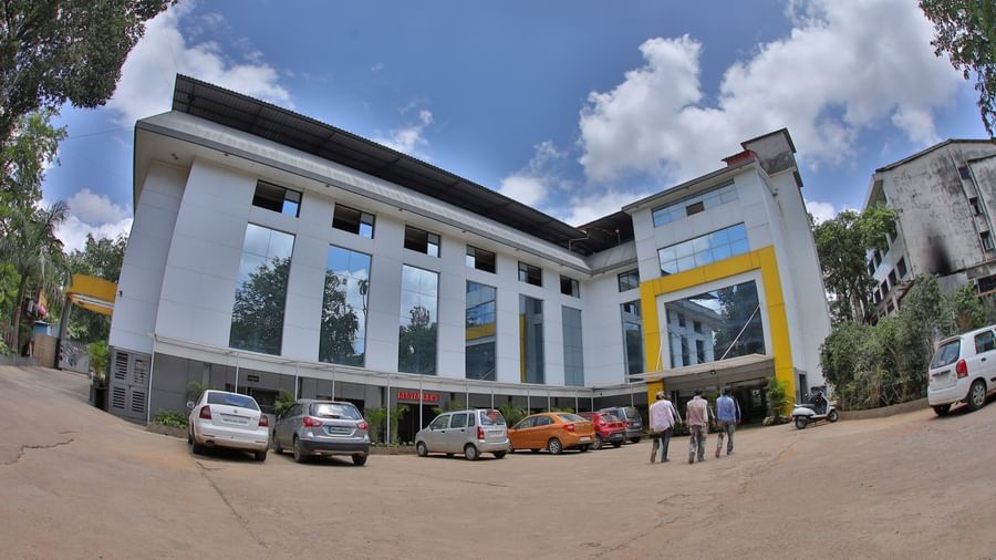The facade of Krushnai Resort building with reflective windows and yellow accents under a partly cloudy blue sky.