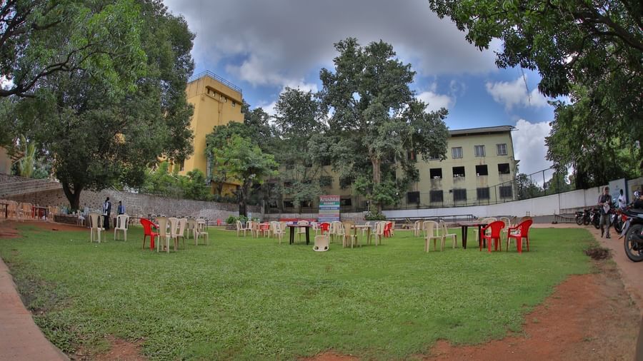 Green lawn at Krushnai Resort with scattered chairs and tables under a cloudy sky, surrounded by tall trees and distant buildings.