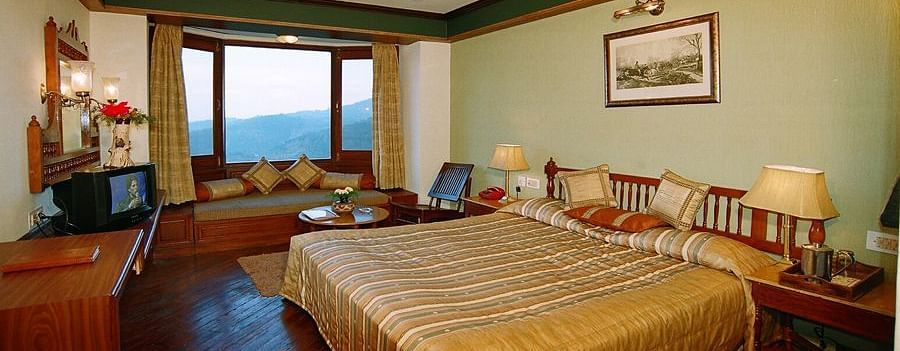 A wide view of a bedroom with a large bed and two side lamps on each side of the bed, a desk and a curved sofa - Sun N Snow Inn, Kausani