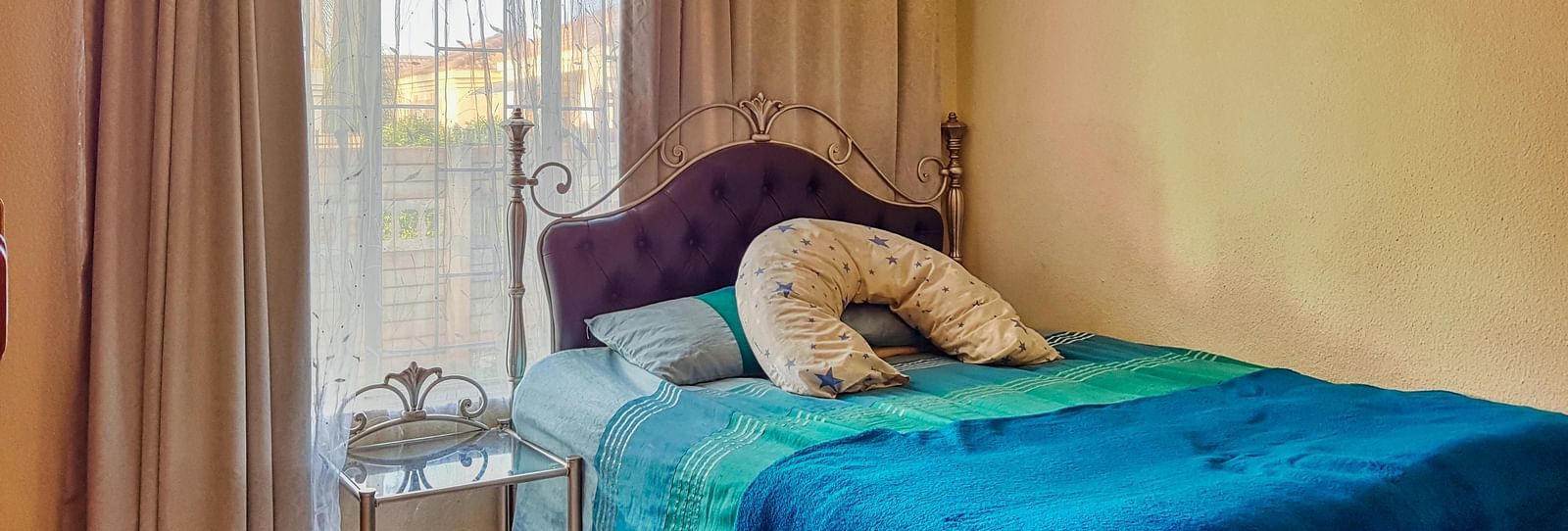 A bedroom featuring a double bed with blue sheets spread neatly over it with beige curtains covering the window behind it.