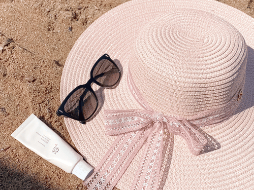Sunscreen, a cap, and sunglasses displayed on sand during a sunny day.