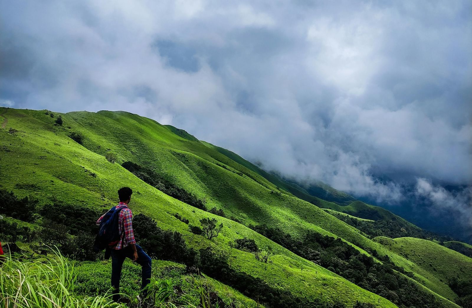  A hiker with a red backpack stands on a grassy trail, looking out at a lush green mountain range under a cloudy sky.