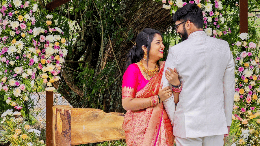 A bride and groom under a floral archway, with the woman in a pink sari looking at the man in a white suit, in an outdoor setting.
