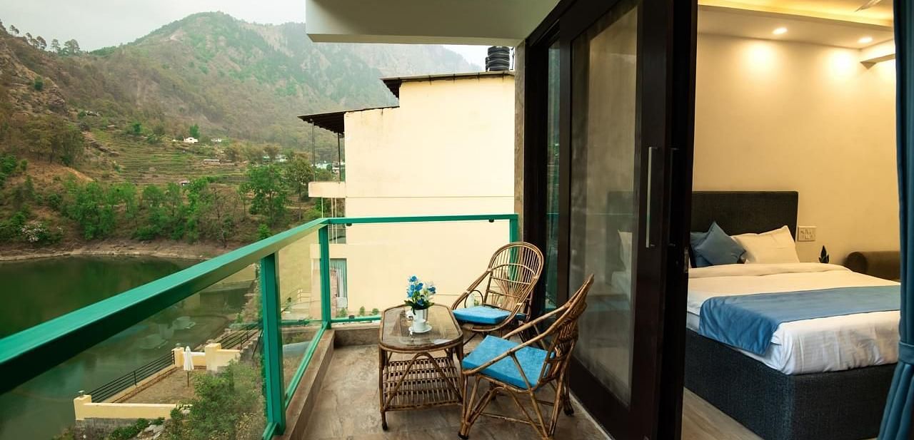 A bedroom with an attached balcony featuring two chairs and a table, overlooking green hills and a scenic mountain view.