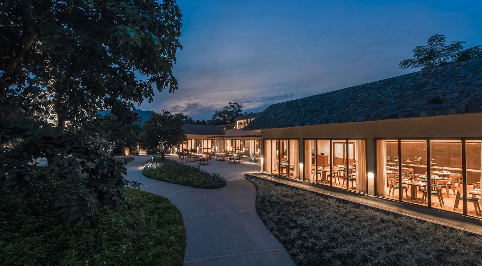 Exterior of a well-lit restaurant at Saraca  Resort & Spa Corbett with mountains in the backdrop during the blue hour