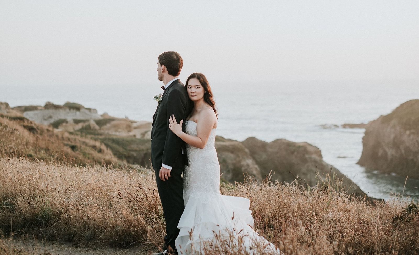 Bride and groom embracing each other on a grassy hill by the ocean