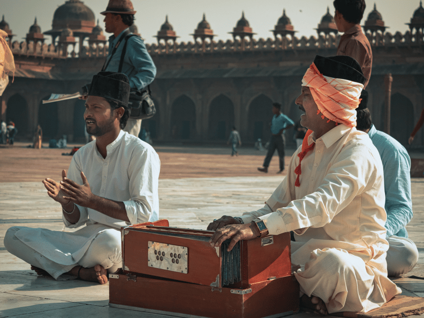 2 men perform traditional music outdoors in a courtyard, with one playing a harmonium and another clapping along.