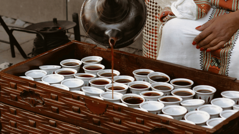 A person pouring coffee to numerous cups, kept inside a small table like structure in the outdoors