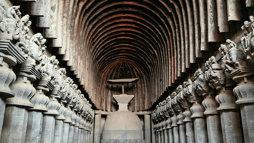 An overview of the prayer hall inside one of the Karla Caves with arches and a giant bell in the distance. These caves are one of the most popular Khandala hill station tourist places.