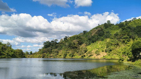 A peaceful lake with lily pads in the foreground, bordered by a lush green hill under a bright blue, cloudy sky.