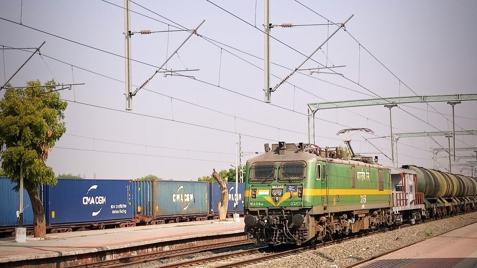 Green and blue passenger train on tracks, with overhead power lines, at a station.