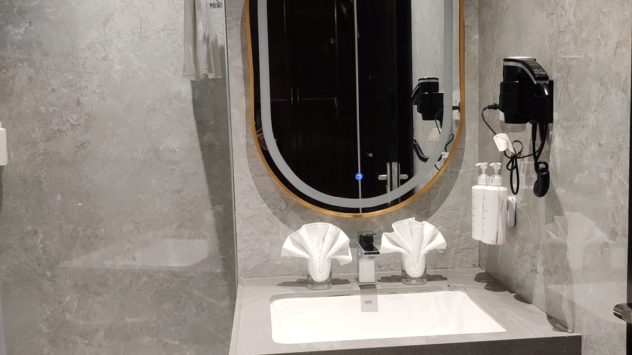 A bathroom at Anant Mahal, Jaipur with a stone vanity, an oval mirror, a wall mounted hair dryer, and a glass shower door.