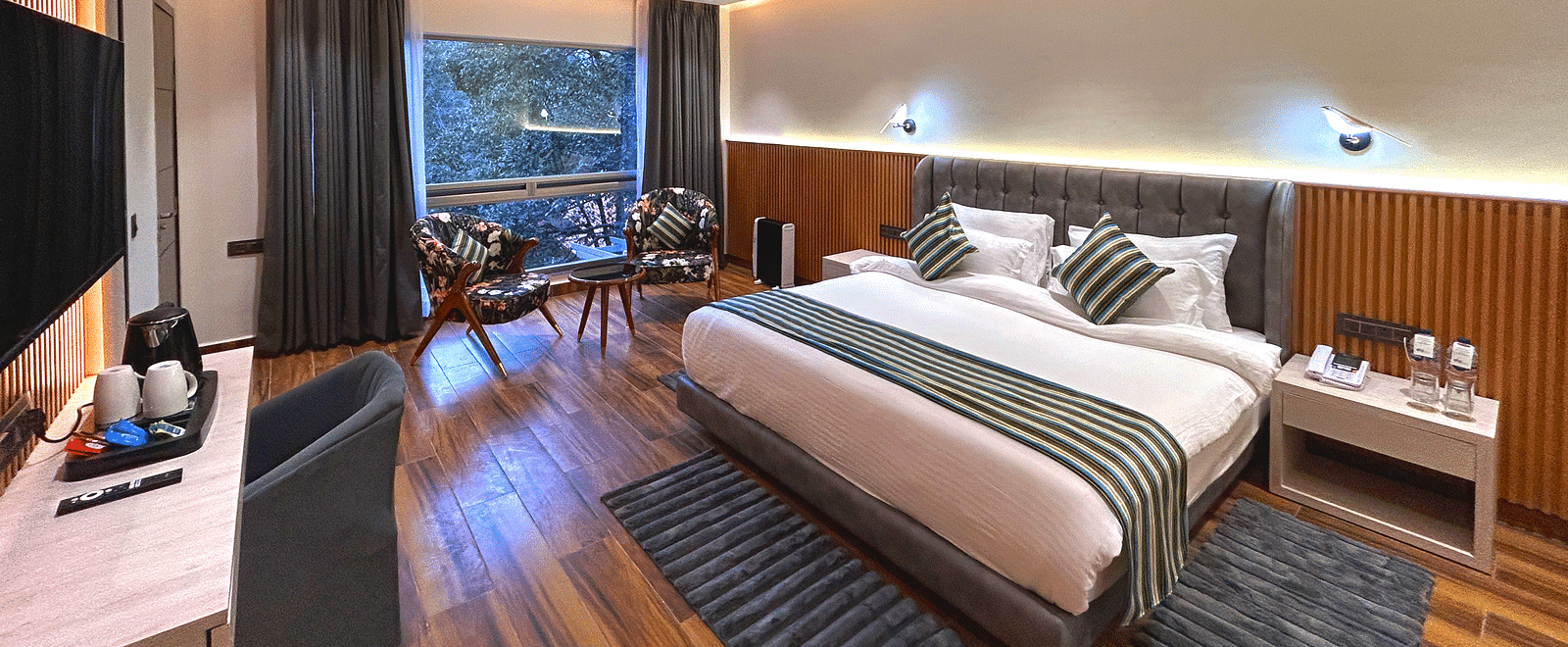 The premium room of DLS The Emerald, Nainital with a bed, bed side table, chairs, coffee table, a window overlooking the view, with warm lighting