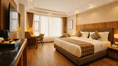 A bedroom at Pride Premier Darjeeling featuring a double size bed, a flat TV screen on the wall, a workstation with chair and desk, a sofa chair, and a large window with curtains drawn.