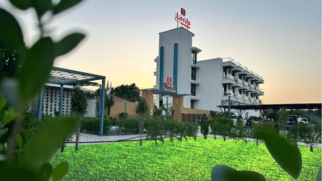 A modern hotel building is seen beyond a green lawn, with plants in the foreground and a clear sky above - Lords SKD Resort, Salangpur