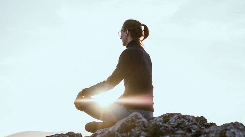 A person sits cross-legged on a rocky surface, meditating with the sun's rays creating a serene glow.