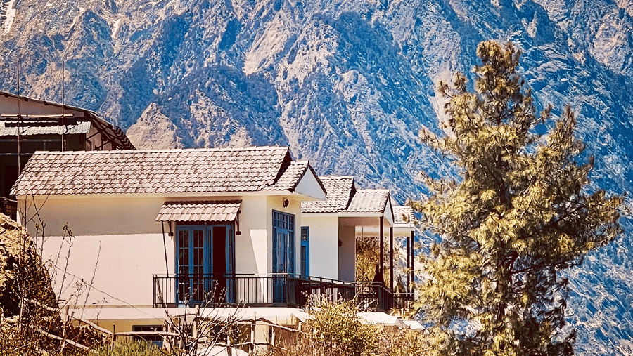A row of cottages with dark windows is situated on a hillside, overlooking a vast, rugged mountain range under a clear blue sky at the location of Faraway Cottages, Auli.