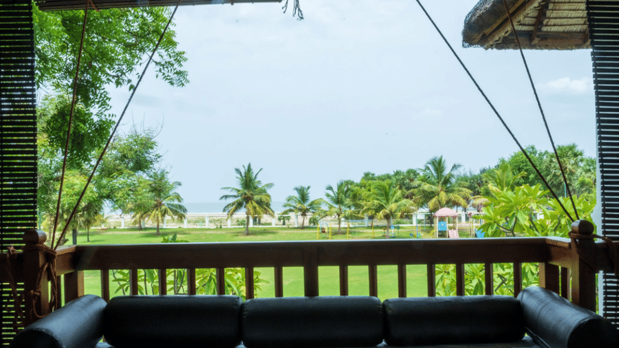 A view from a covered wooden veranda overlooking a green lawn with trees under a clear sky | Le Pondy
