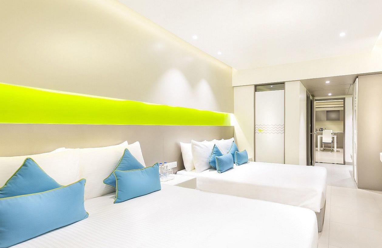A spacious Suite features a large bed with a headboard stretching across the wall. Cushion chairs sit beside a wooden bedside table, and a wall-mounted television is positioned opposite the bed