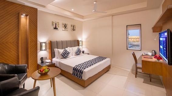 A comfortable hotel bedroom featuring a double bed with white linens and patterned cushions, a wooden bedside table with a lamp and fruit bowl, a black armchair, a writing desk with a chair, and a television on the wall at Lords Inn Motikhavdi, Jamnagar.