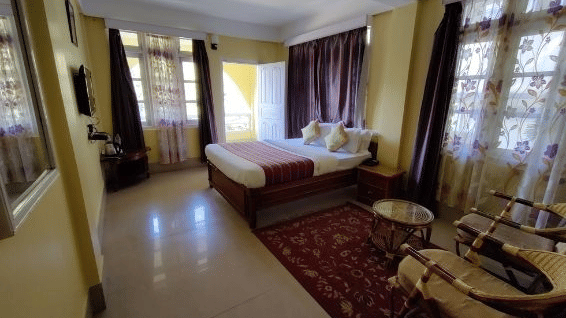 A side view of a double bed room with marble flooring, tables, carpet, door and windows with curtains