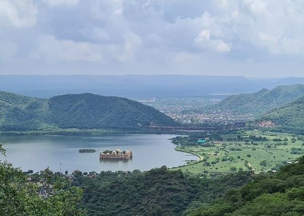 A palace appears to float in a lake, surrounded by lush hills and a distant town, under a cloudy sky.