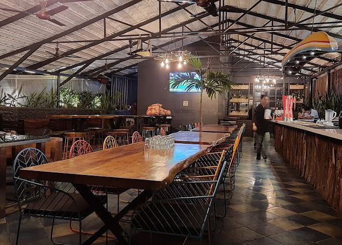  Indoor restaurant space with long wooden tables and chairs, under an exposed ceiling with hanging lights | Astra Hotels and Suites Kadubeesanahalli