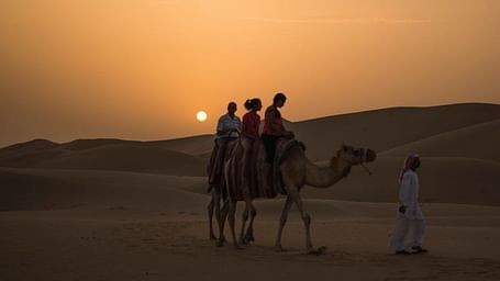 image of three people enjoying a camel back ride with a beautiful sunset in the background with sand dunes .