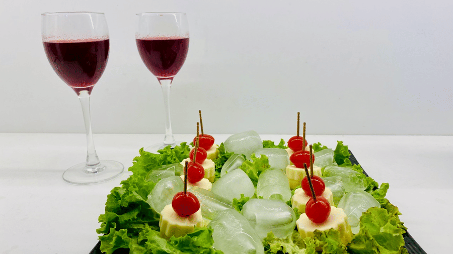 Black tray of cheese, cherry and pineapple cocktail picks on lettuce with crushed ice, served beside 2 glasses of red wine on a white surface.
