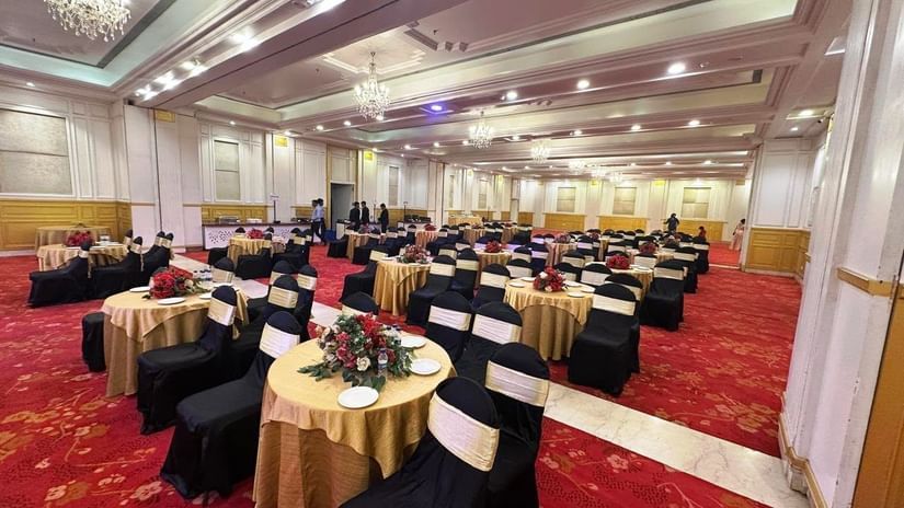  A banquet hall at Amer Greens is set for an event with cocktail tables draped in gold linen, surrounded by chairs in black covers and gold sashes, on a red and gold patterned carpet.